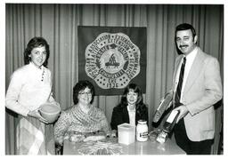 Four People Posing with Food and Michigan Cooperative Association Extension Homemakers Service Sign (Part of the NMU Historic Photographs Collection)